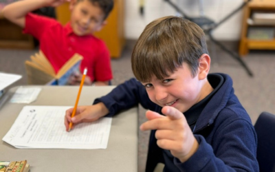 Young boy at a desk points at the camera while another boy reads in the background at Redeemer Lutheran School.