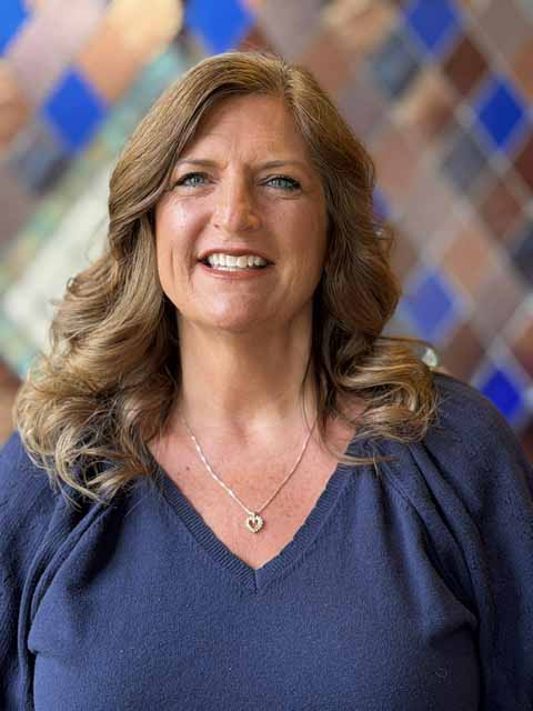 Debbie Grecu, a smiling woman with long brown hair, wearing a blue sweater and a heart necklace.