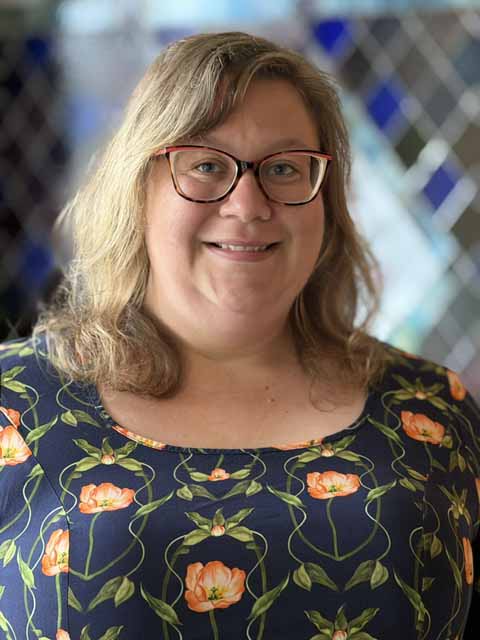 Emily Farrell, a woman with blonde hair and glasses, smiles in a floral dress.