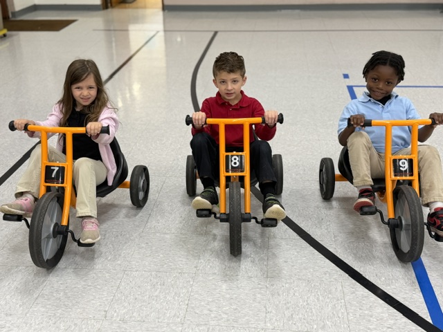 Three children on tricycles in a school gym, numbers 7, 8, 9 visible