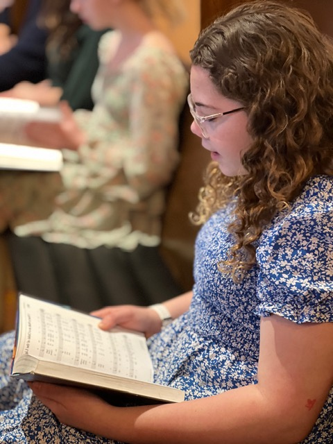 Young woman in glasses and blue dress reads from hymnal at Redeemer Lutheran School