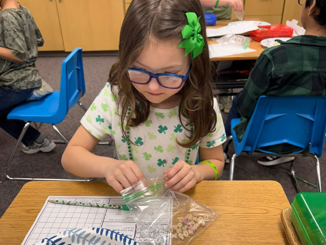 Young girl with blue glasses and green bow works on a school assignment.