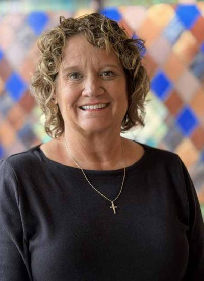 Angie Plutat, a woman with curly blonde hair, smiles in front of a colorful background.