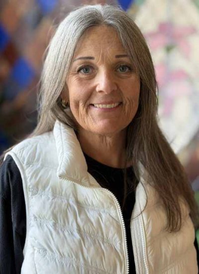 Carrie Cardwell, a woman with long gray hair and a white vest, smiles warmly.