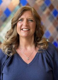 Debbie Grecu, a smiling woman with long brown hair, wearing a blue sweater and a heart necklace.