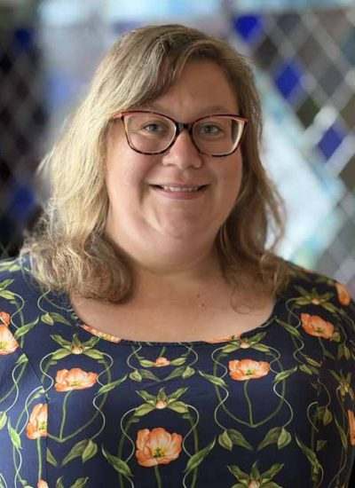 Emily Farrell, a woman with blonde hair and glasses, smiles in a floral dress.