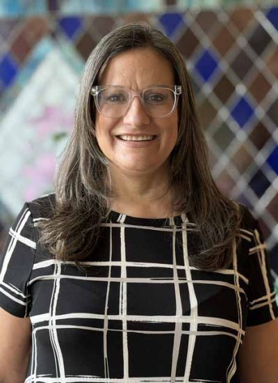 Laura Collins, a woman with long, graying hair and glasses, smiles in a black and white plaid top.