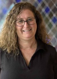 Sallie Miller, a smiling woman with curly brown hair and glasses, wearing a black shirt.